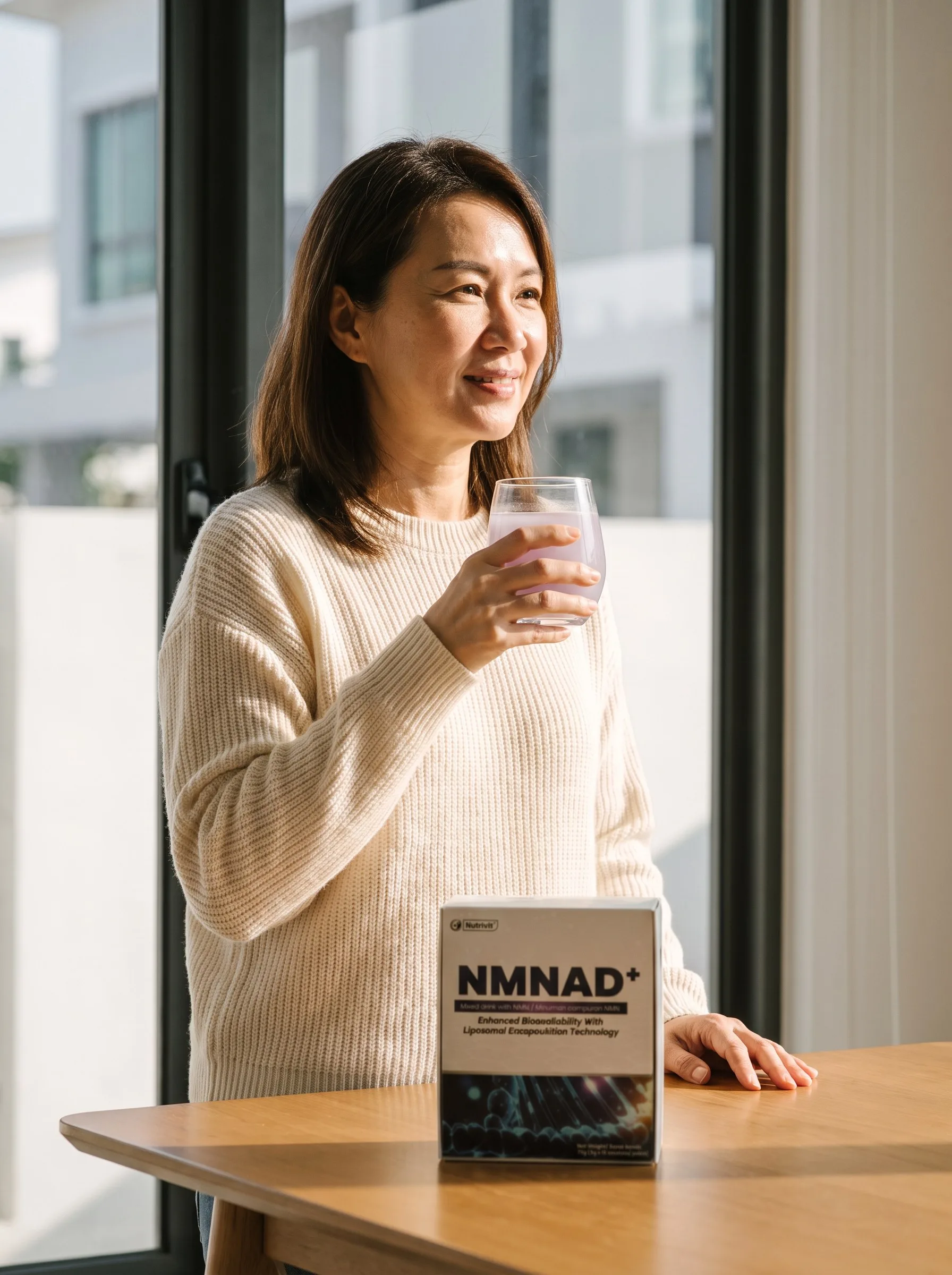 A woman by a sunlit window holding a glass, NMNAD+ on the table